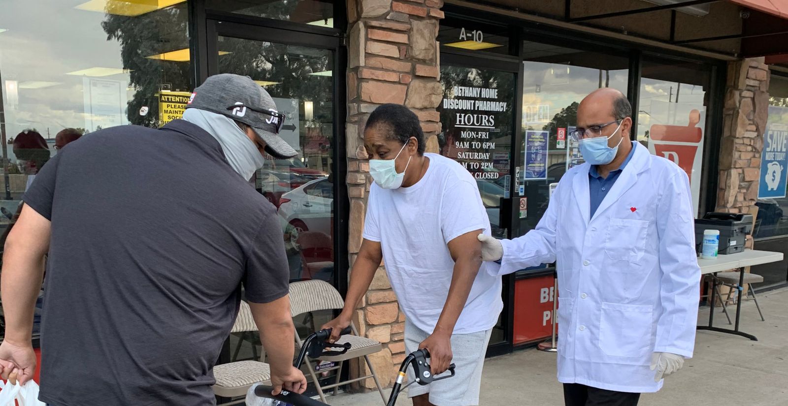 A pharmacist in a white coat and mask helps a woman using a walker