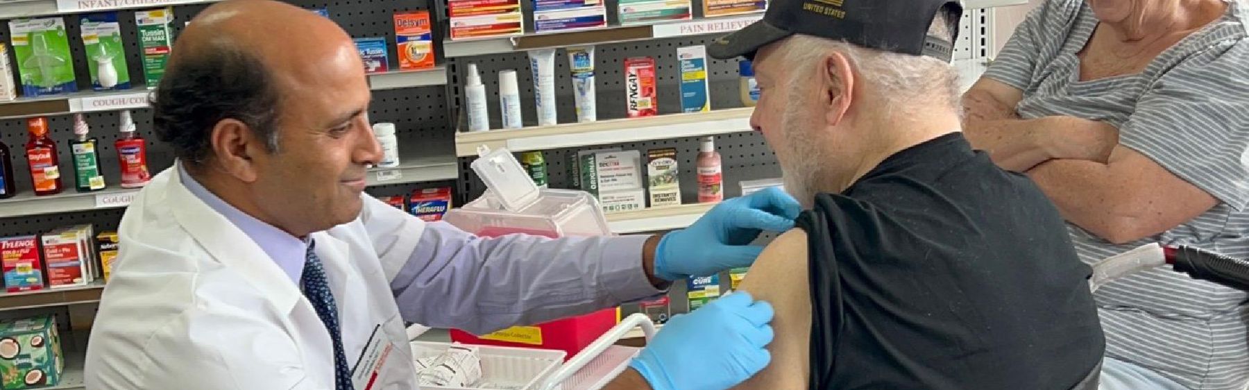 A pharmacist in a white coat and gloves gives a vaccine to an older man in a baseball cap